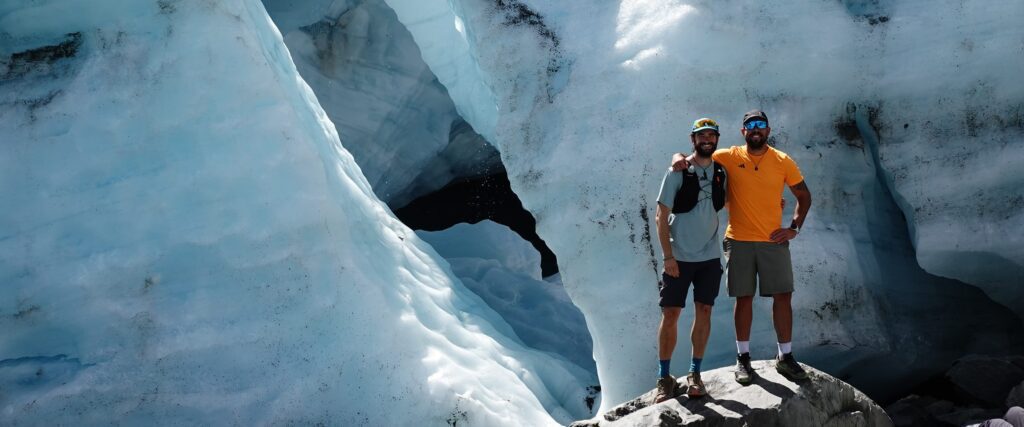 2 personas sobre el hielo de un glaciar, junto a una cueva de hielo